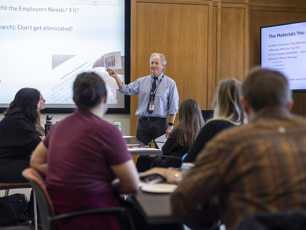 A man in a blue shirt holds up a chart in front of a group of students.