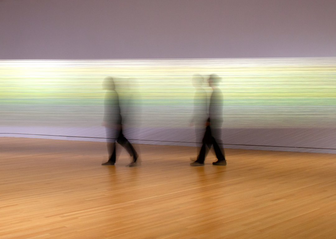 Two blurred figures in black walk in front of an art installation of bright threads strung between two walls.