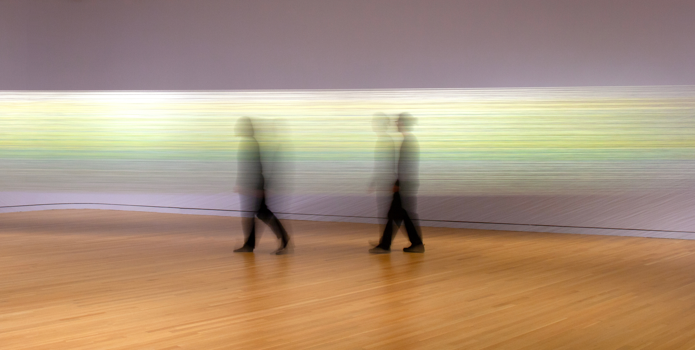 Two blurred figures in black walk in front of an art installation of bright threads strung between two walls.