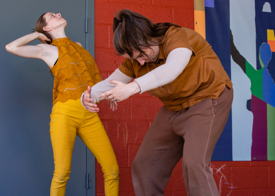 Two dancers, a man and a woman in yellow pants and top, pose mid-movement against a red cement wall. 