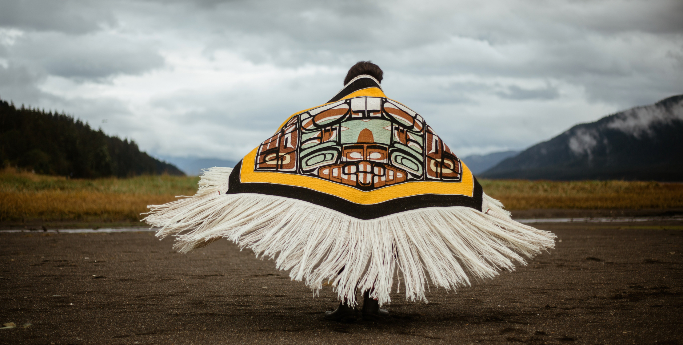 A woman with her back to the camera wears a floor-length fringed blanket wrapped around her shoulders in a cloudy landscape. 