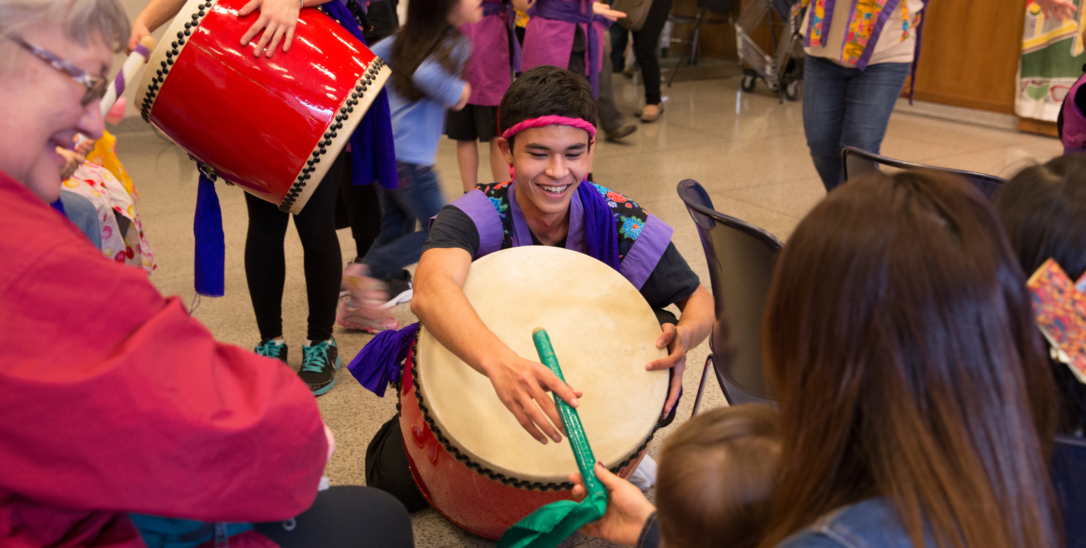 A young male dancer sits on the floor holding a large drum