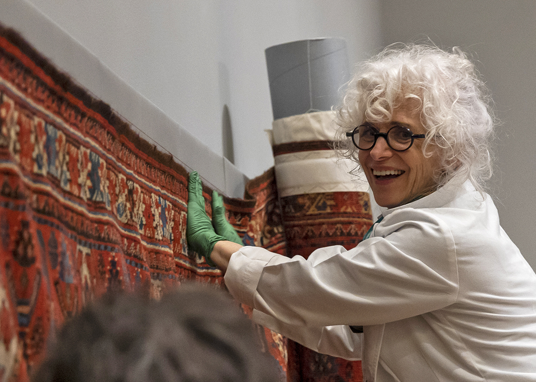A smiling conservator helps hang a prayer rug on a gallery wall