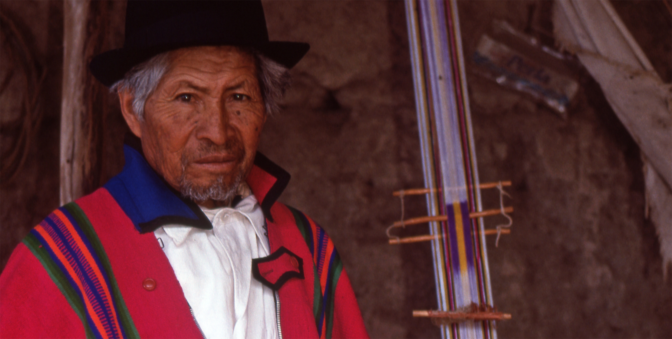 An Ecadudorian weaver in a black hat and traditional red wrap stands next to his loom