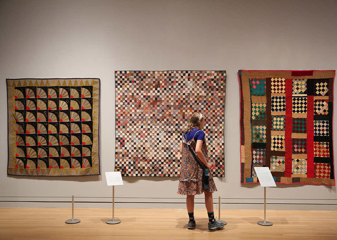 A visitor walks past three colorful quilts hanging on a gallery wall