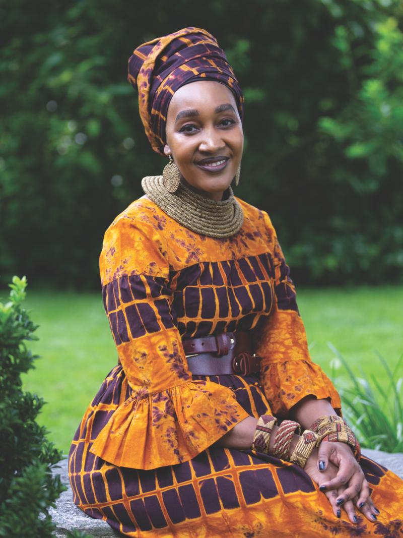 Woman wearing a bright orange and purple dress and head wrap seated on a stone wall