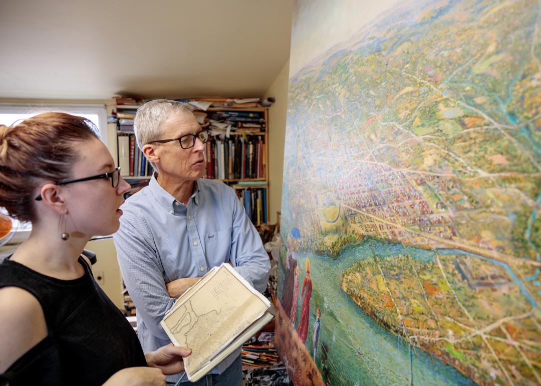 A young brunette woman and a man with a blue shirt and white hair look at a large bird's-eye view painting.