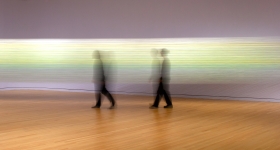 Two blurred figures in black walk in front of an art installation of bright threads strung between two walls.