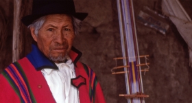 An Ecadudorian weaver in a black hat and traditional red wrap stands next to his loom